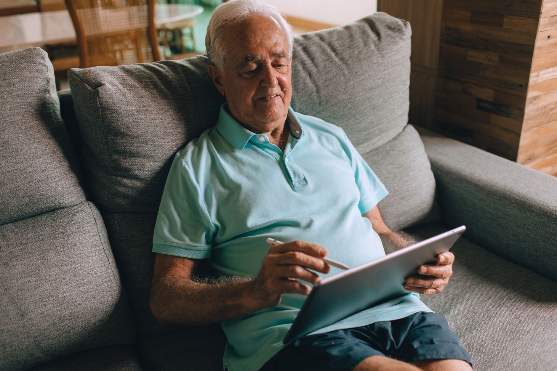 Man sitting on a couch using a tablet with a stylus indoors — the kind of relaxed, from-home setup this remote admin work allows.